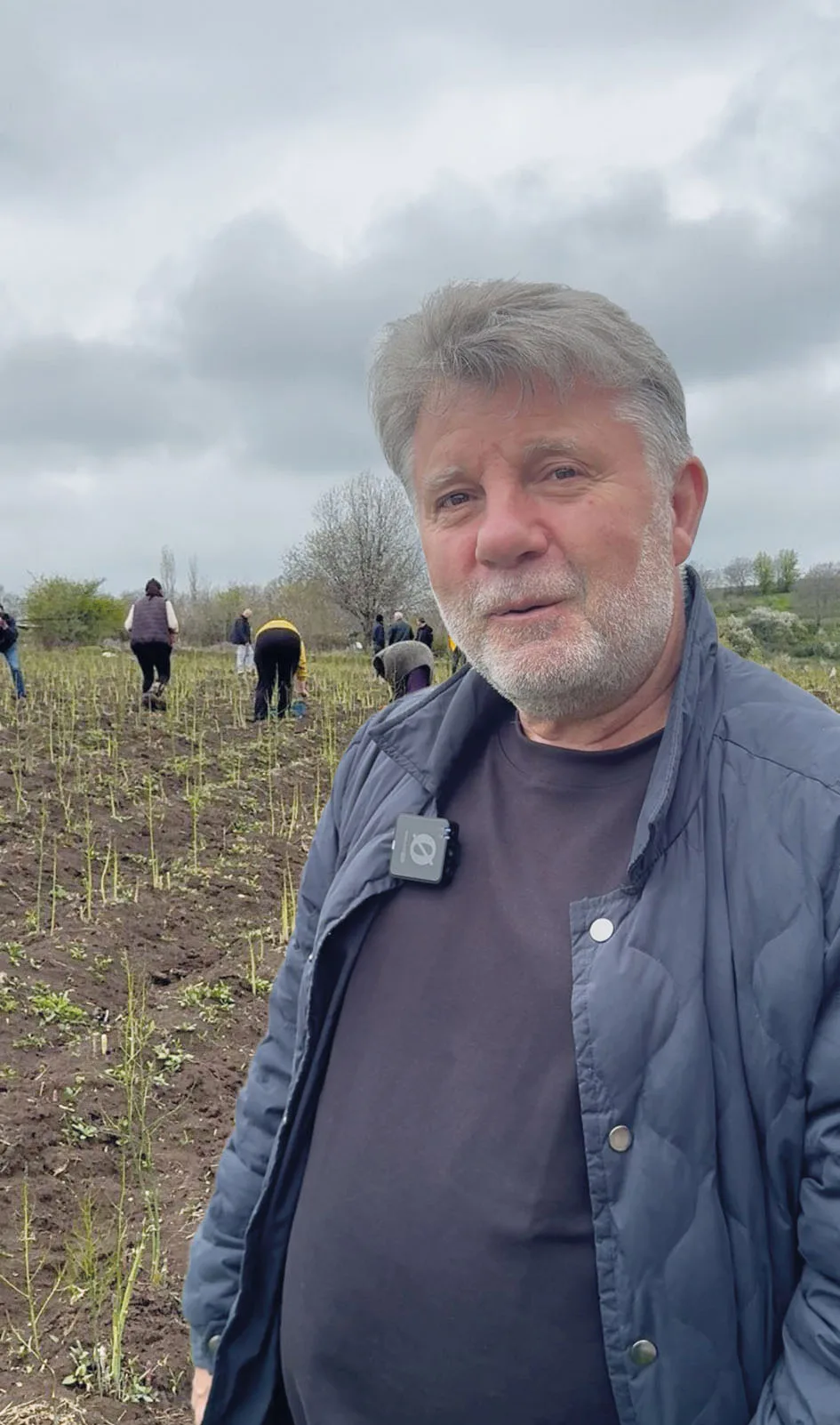 Lionel Balian, veste bleue, debout dans un champ d’asperges à Kornidzor, des femmes récoltant à l’arrière-plan